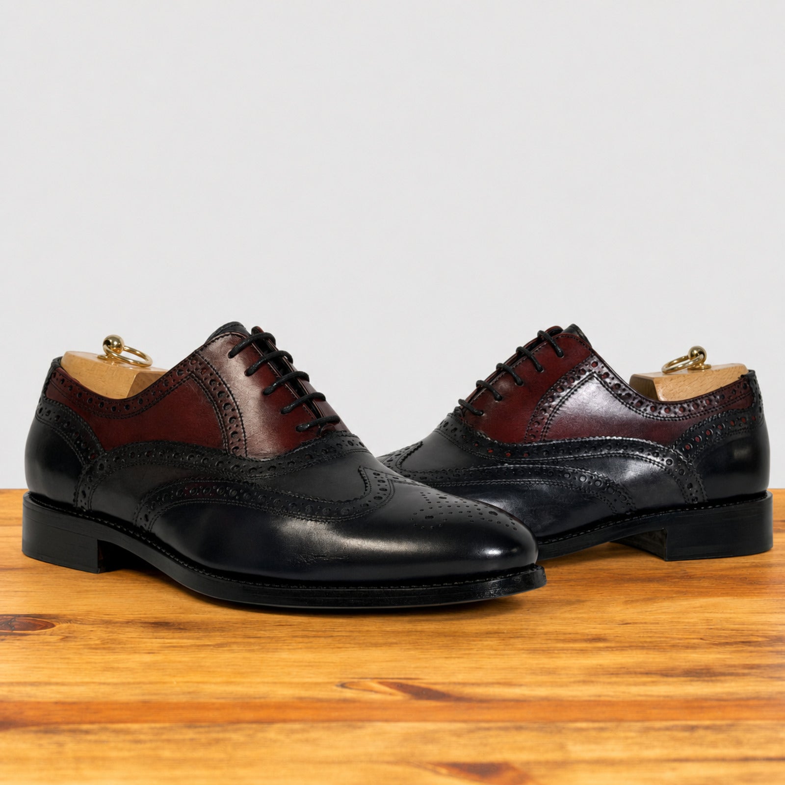A pair of men's wingtip oxford shoes in blue and dark burgundy leather, displayed on a wooden surface against a light gray background.
