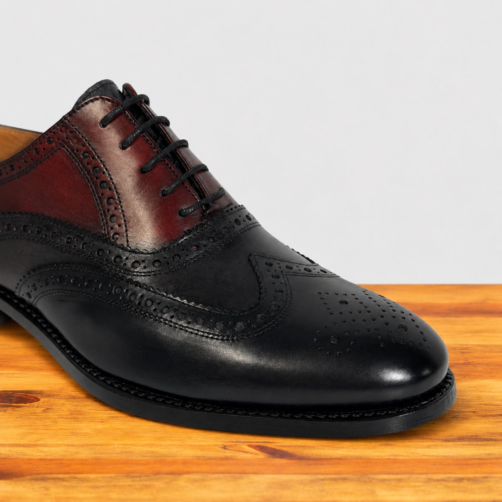 A men's wingtip oxford shoe in blue and dark brown leather, featuring decorative brogue perforations, a black lace, and a black sole, displayed on a wooden surface against a light gray background.