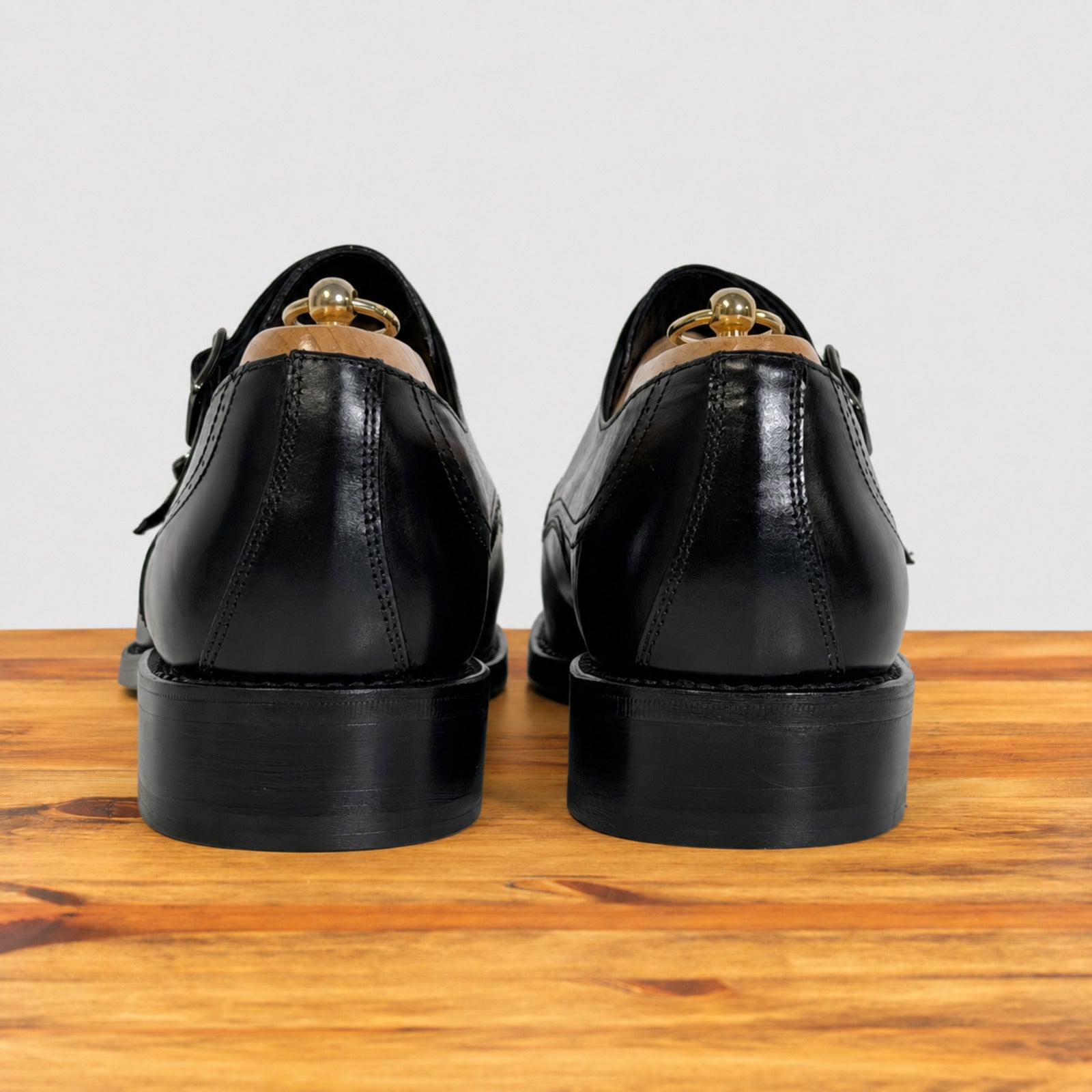 A pair of black leather lace-up dress shoes shown from the back, resting on a wooden surface against a light gray background.
