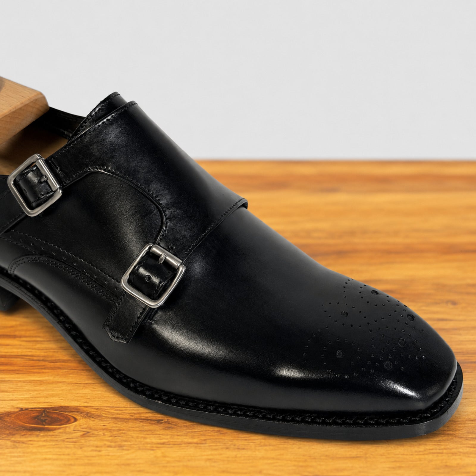 A black leather double strap monk-style dress shoe with silver-tone buckles, shown resting on a wooden surface against a light gray background.