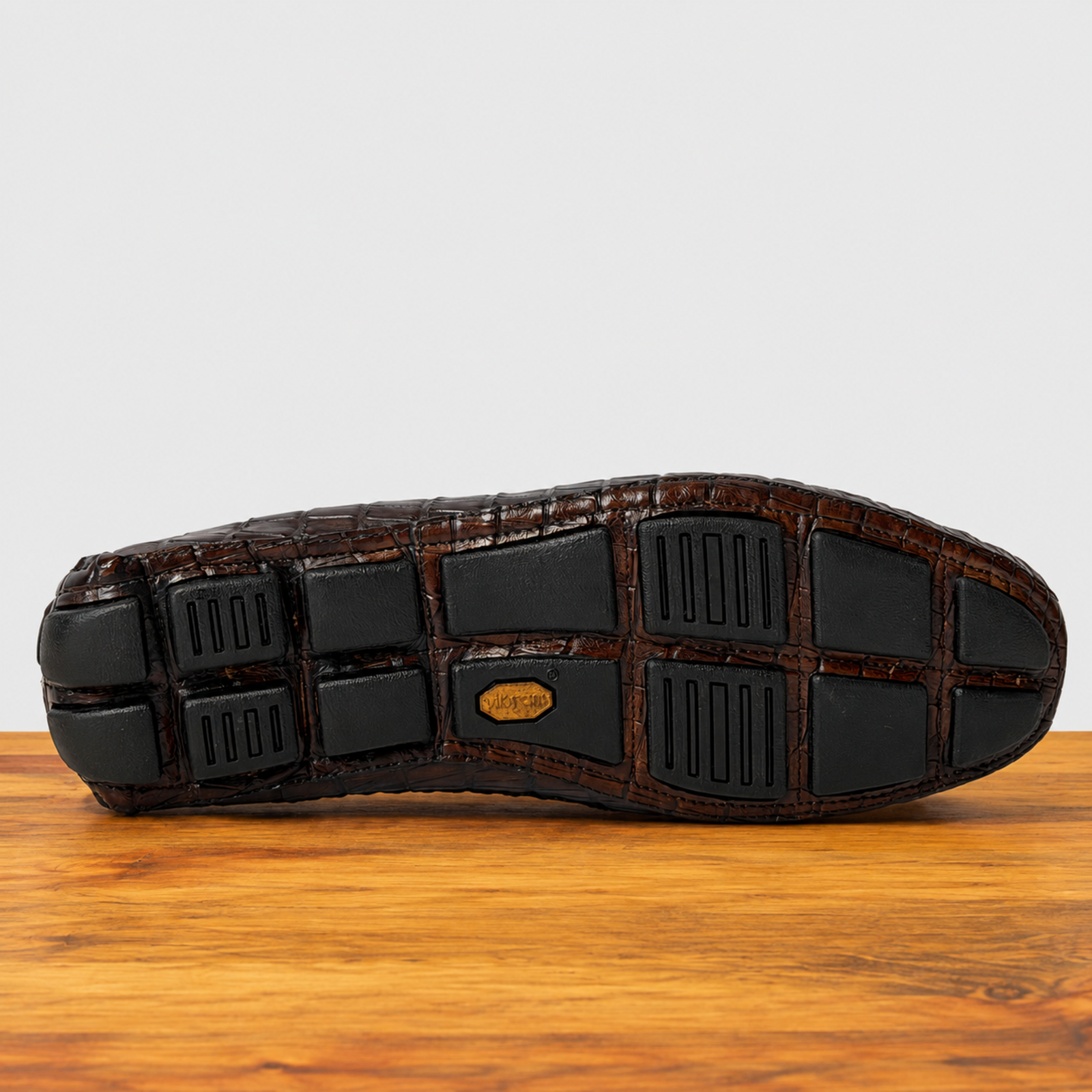 The outsole of a cerris color crocodile- leather shoe, showing black rubber tread blocks and a small gold-tone logo medallion, resting on a wooden surface against a light gray background.