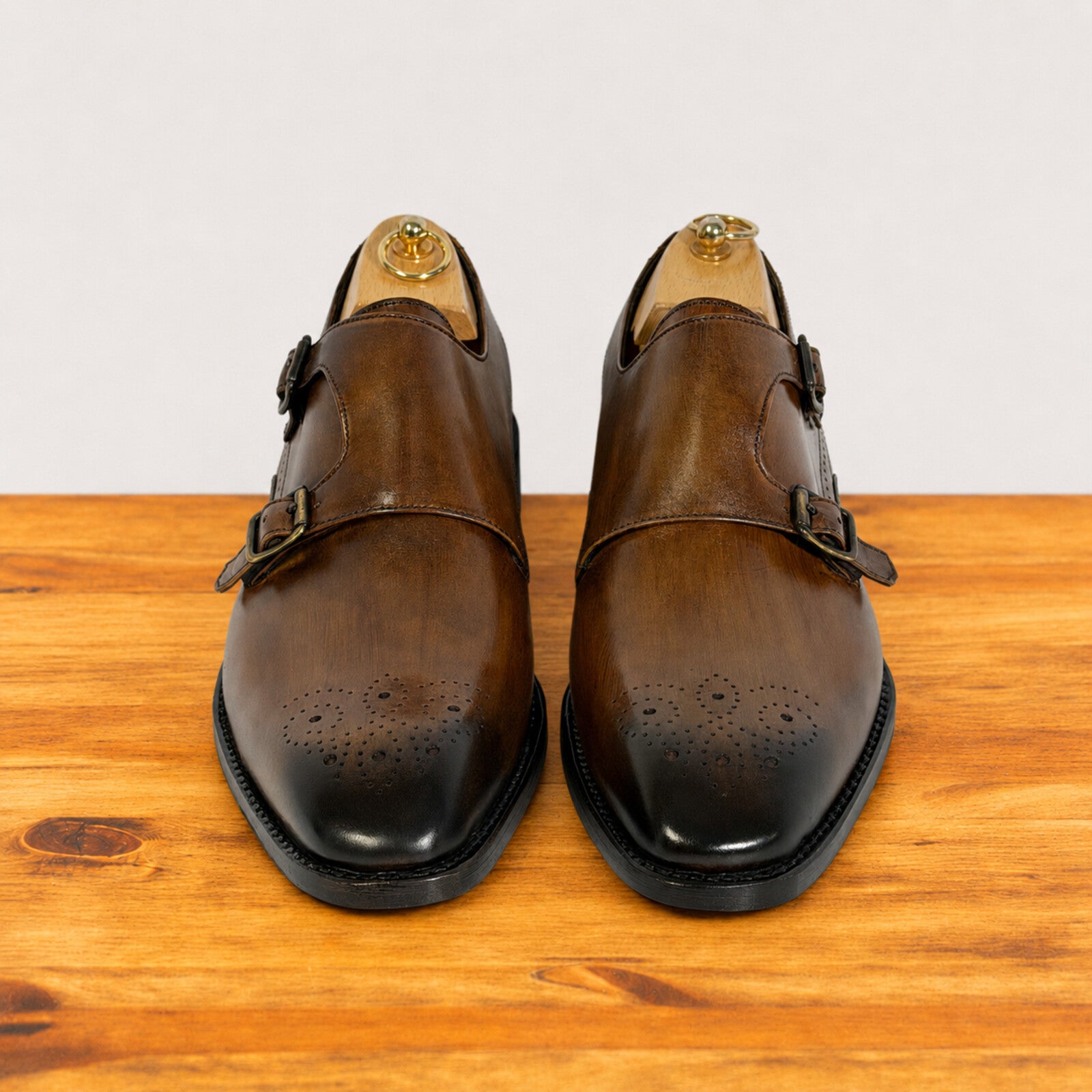 A pair of chestnut leather double monk shoes with brogue detailing on the toes, displayed on wooden shoe trees on a wooden surface against a light gray background.