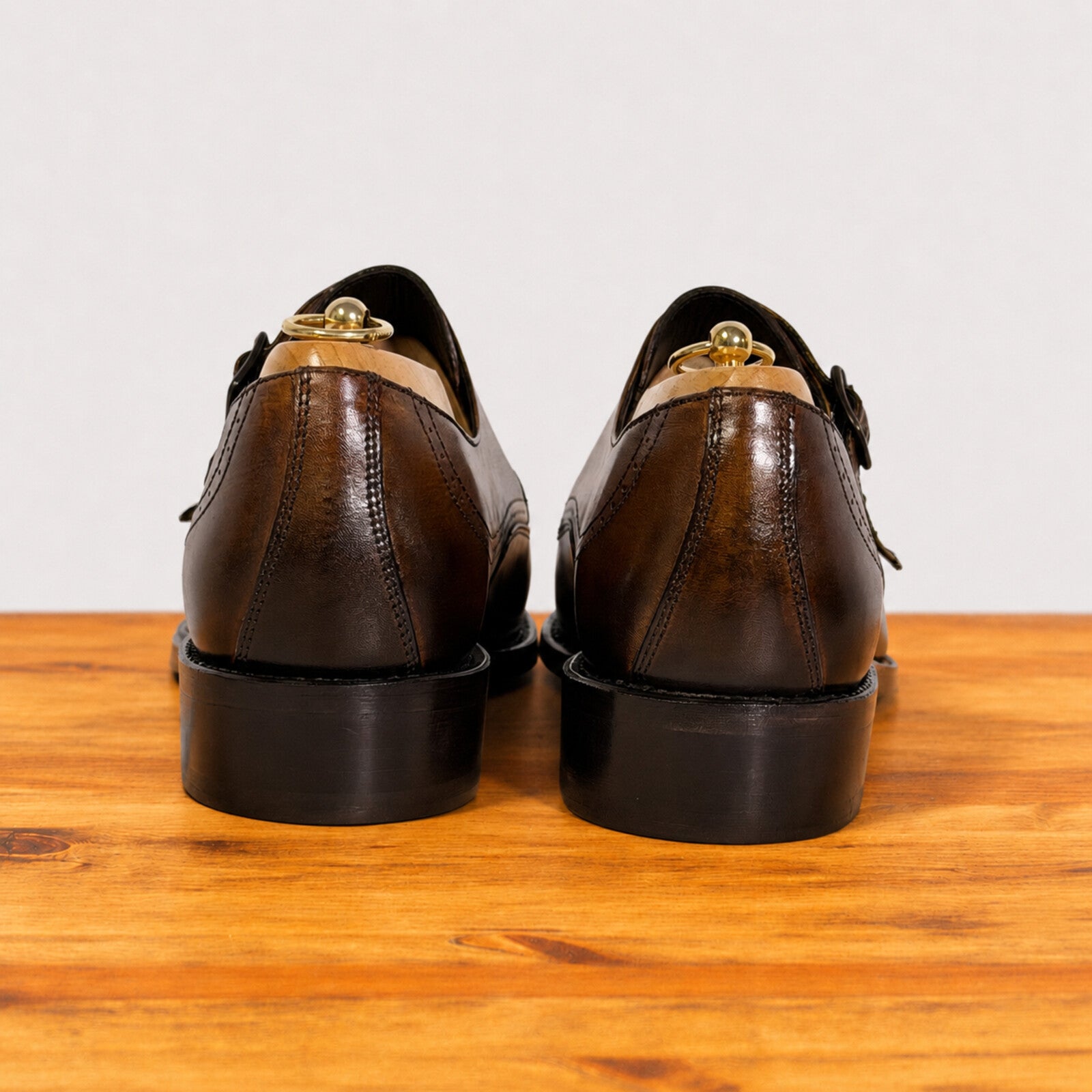 A pair of chestnut leather lace-up shoes shown from the back, resting on a wooden surface against a plain light background.