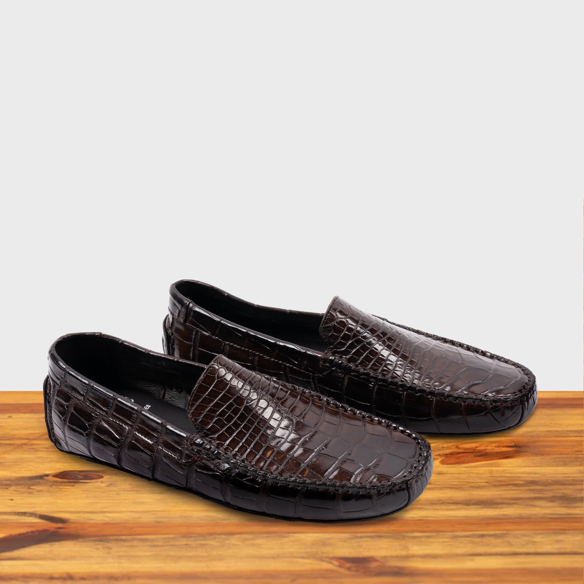 A Pair of Dark Brown crocodile-patterned loafer on a wooden surface with a light gray background