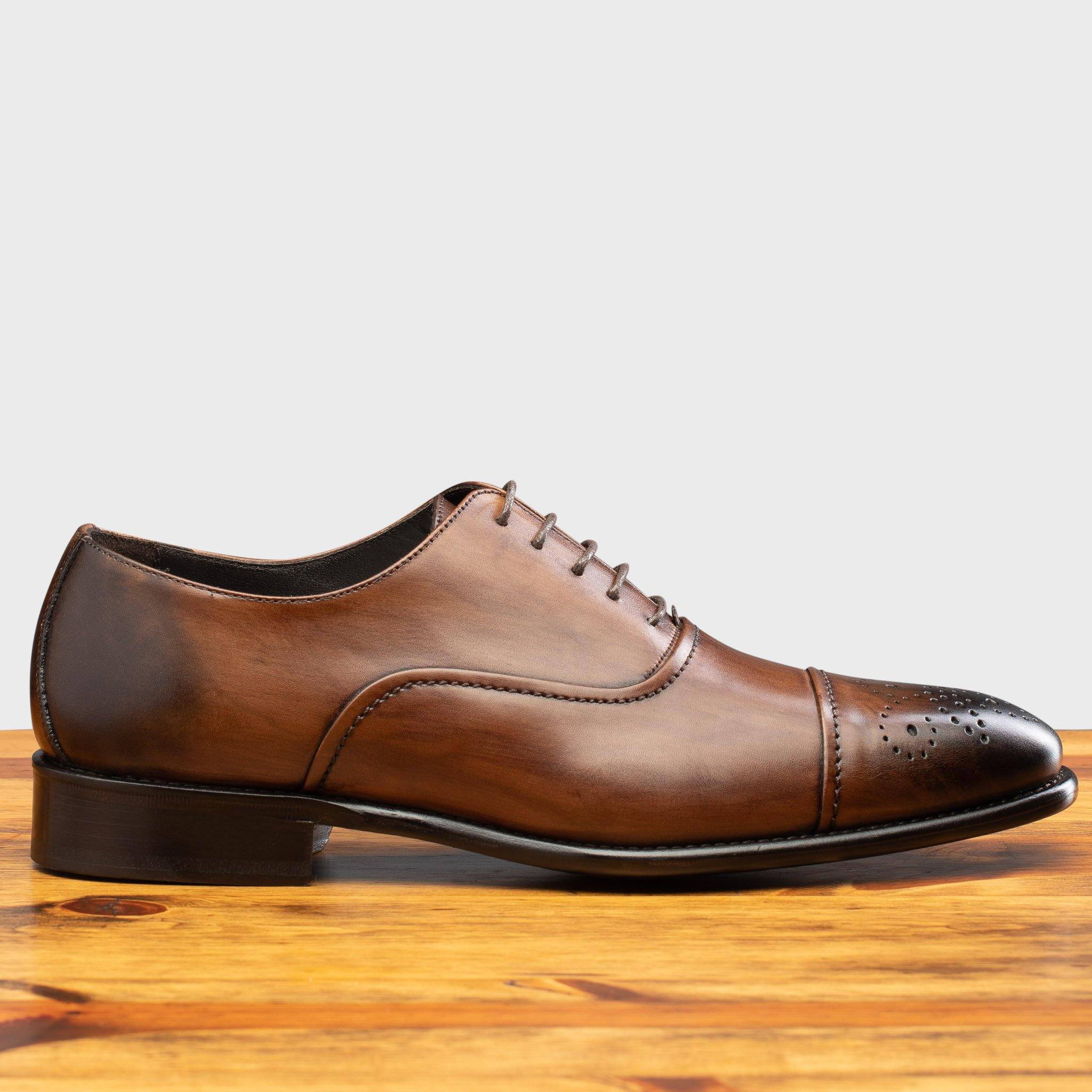 Brown leather oxford dress shoe with a cap toe and subtle perforated detailing, shown in side profile on a wooden surface.