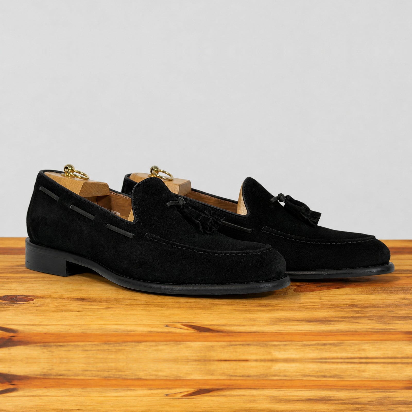 A pair of  black suede tassel loafer shown in profile on a wooden surface against a light gray background.