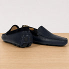 A pair of blue leather driving loafers with a distinctive black rubber sole, displayed on a light wood surface against a white background.