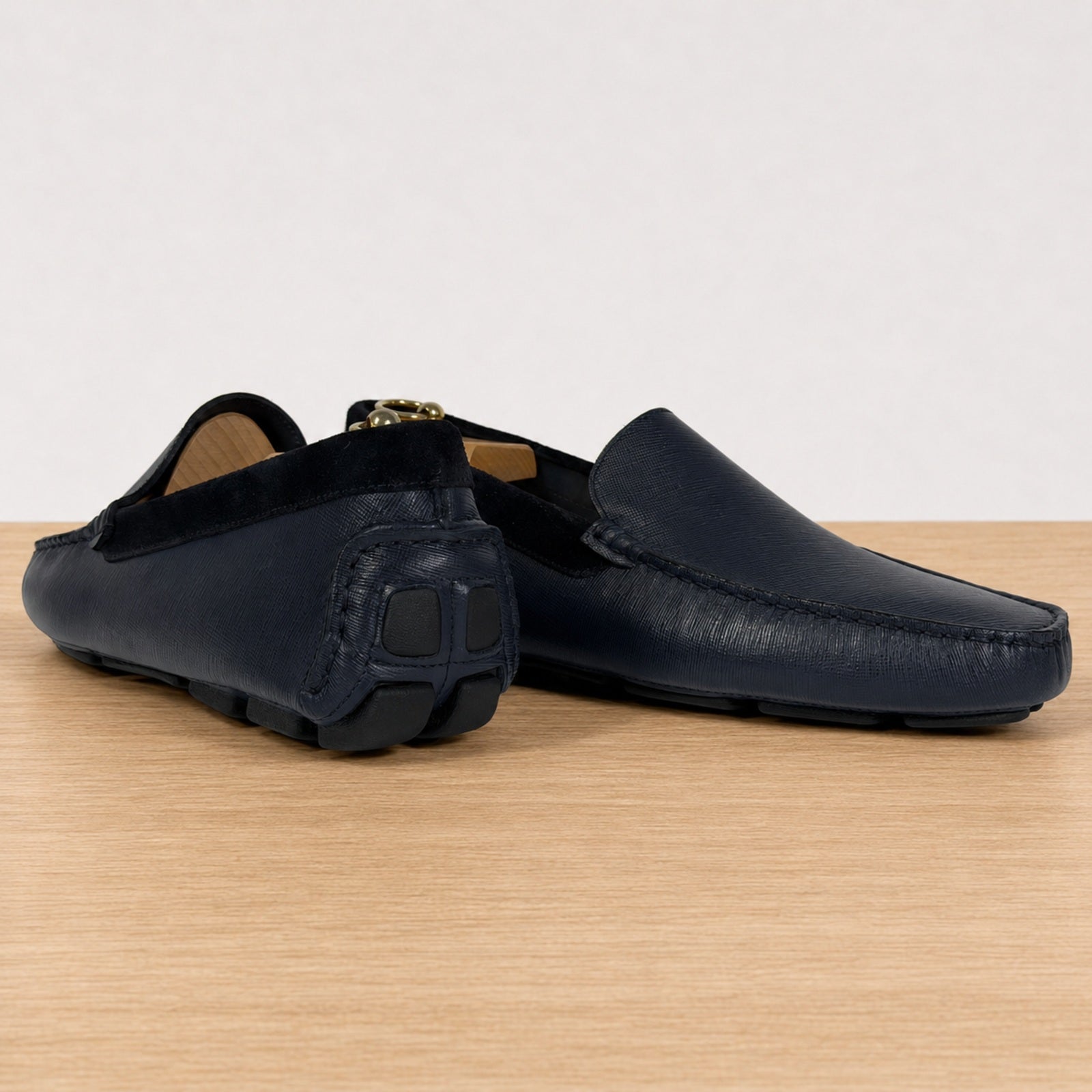 A pair of blue leather driving loafers with a distinctive black rubber sole, displayed on a light wood surface against a white background.