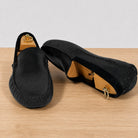 A pair of black slip-on loafers with a textured woven upper, displayed on a light wood surface against a white background.
