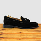A single black suede tassel loafer shown in profile on a wooden surface against a light gray background.