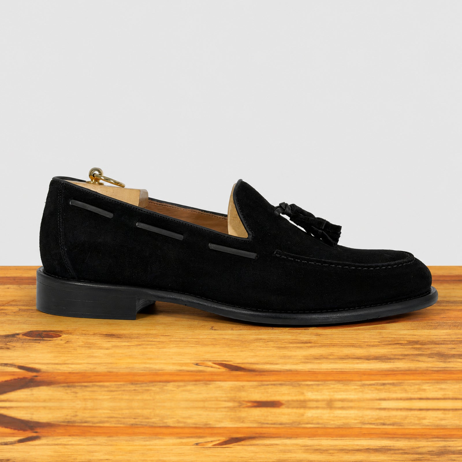 A single black suede tassel loafer shown in profile on a wooden surface against a light gray background.