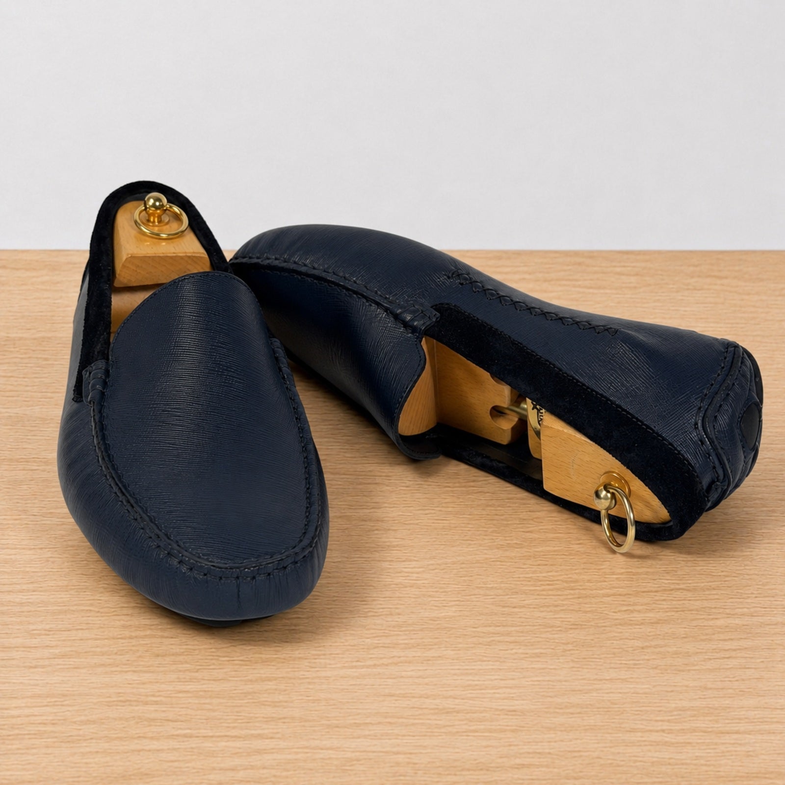 A pair of blue leather slip-on loafers displayed on wooden shoe trees on a light wood tabletop against a white background.