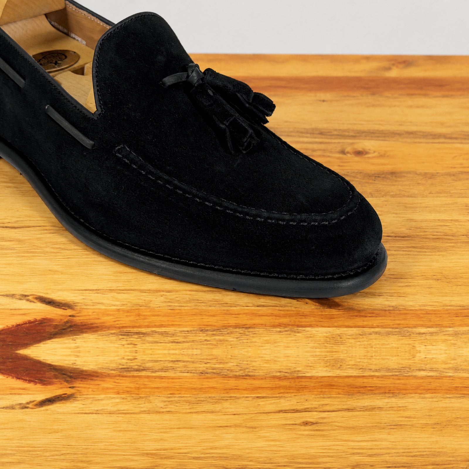 A close up of black suede tassel loafer shown in profile on a wooden surface against a light gray background.