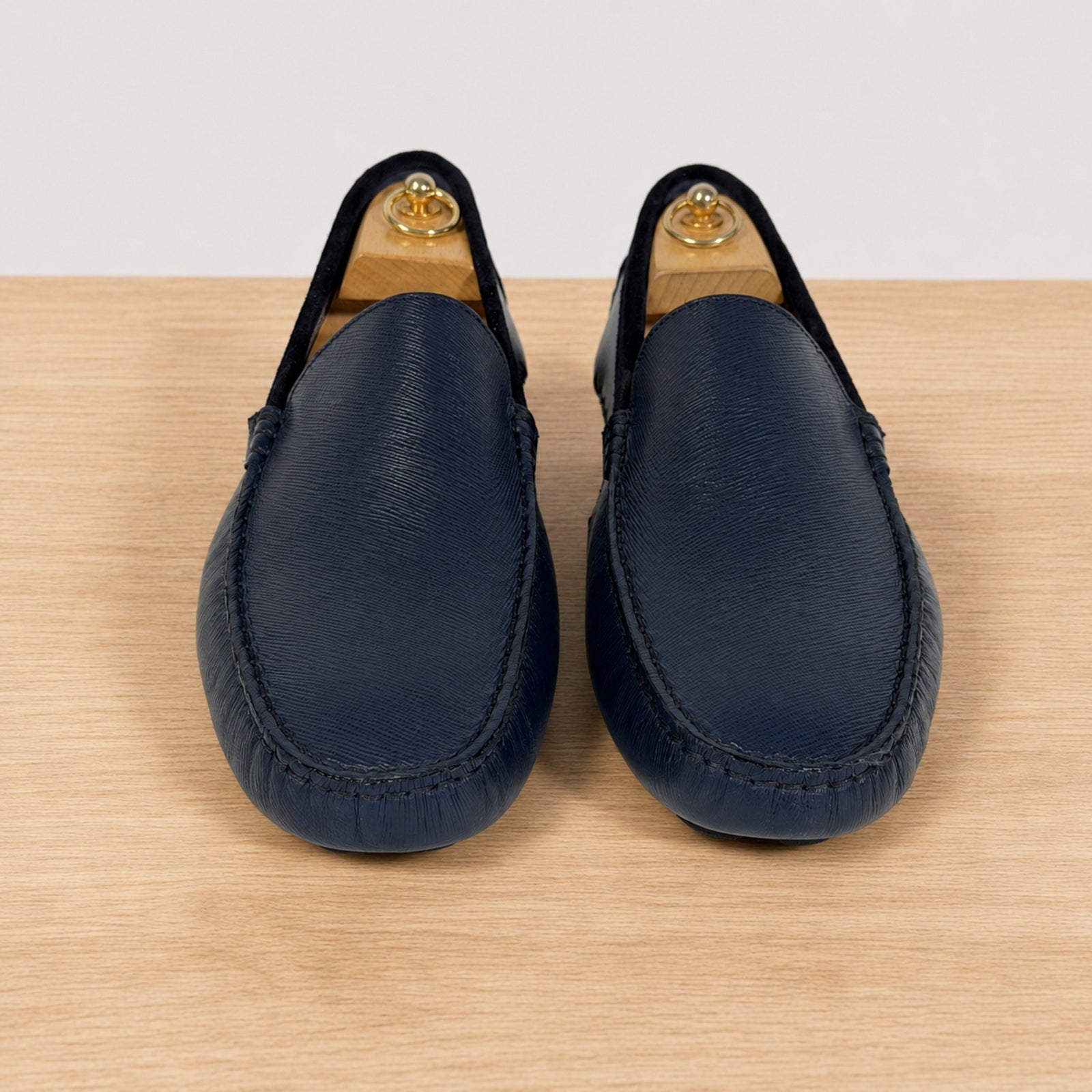 A pair of  blue leather driving loafers with a pebbled texture, displayed on a light wood surface against a neutral light gray background.
