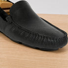 A close-up of a black textured leather driving loafer on a light wood surface.