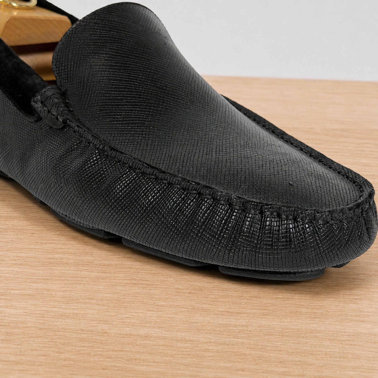 A close-up of a black textured leather driving loafer on a light wood surface.