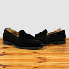 A pair of black suede tassel loafer shown in profile on a wooden surface against a light gray background.