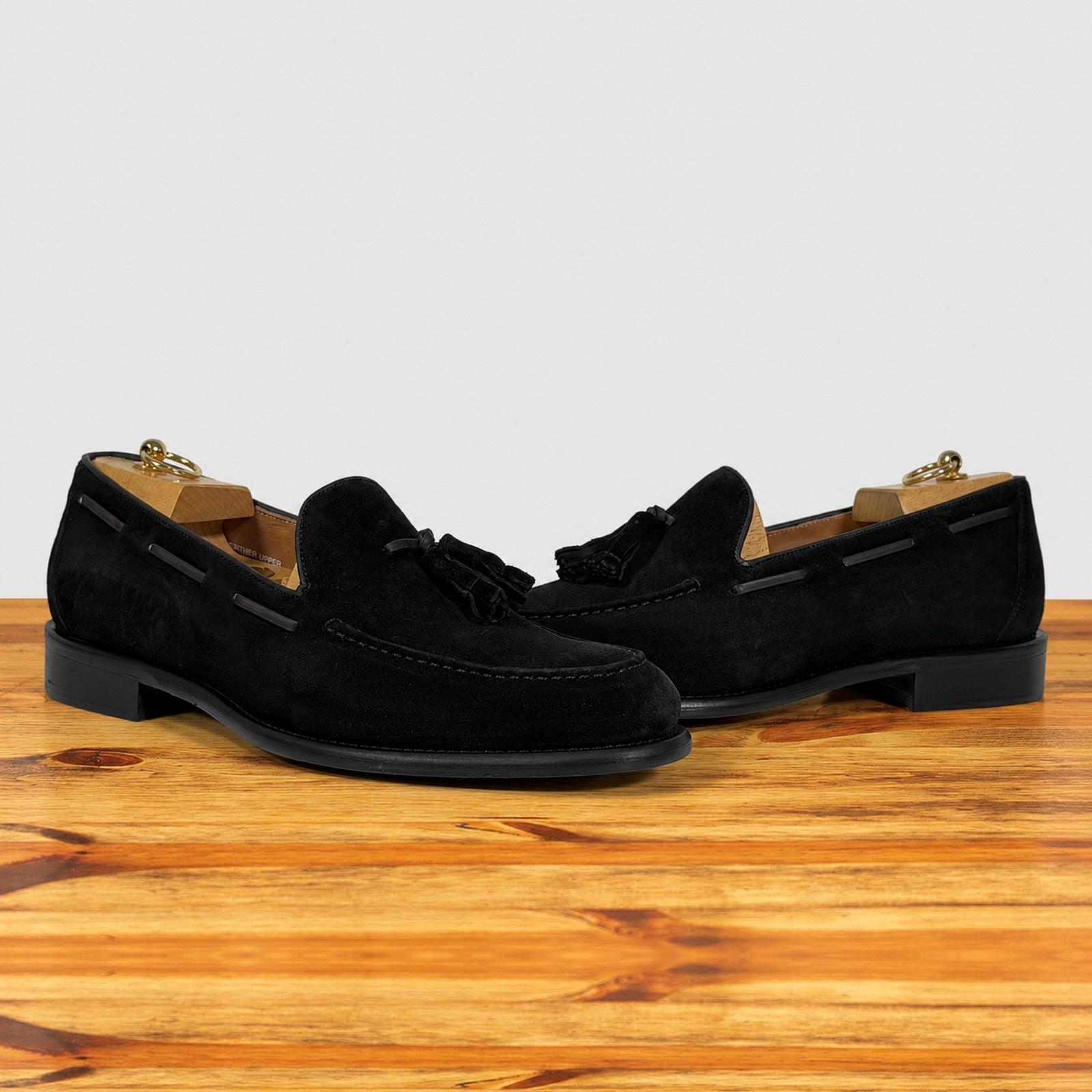 A pair of black suede tassel loafer shown in profile on a wooden surface against a light gray background.