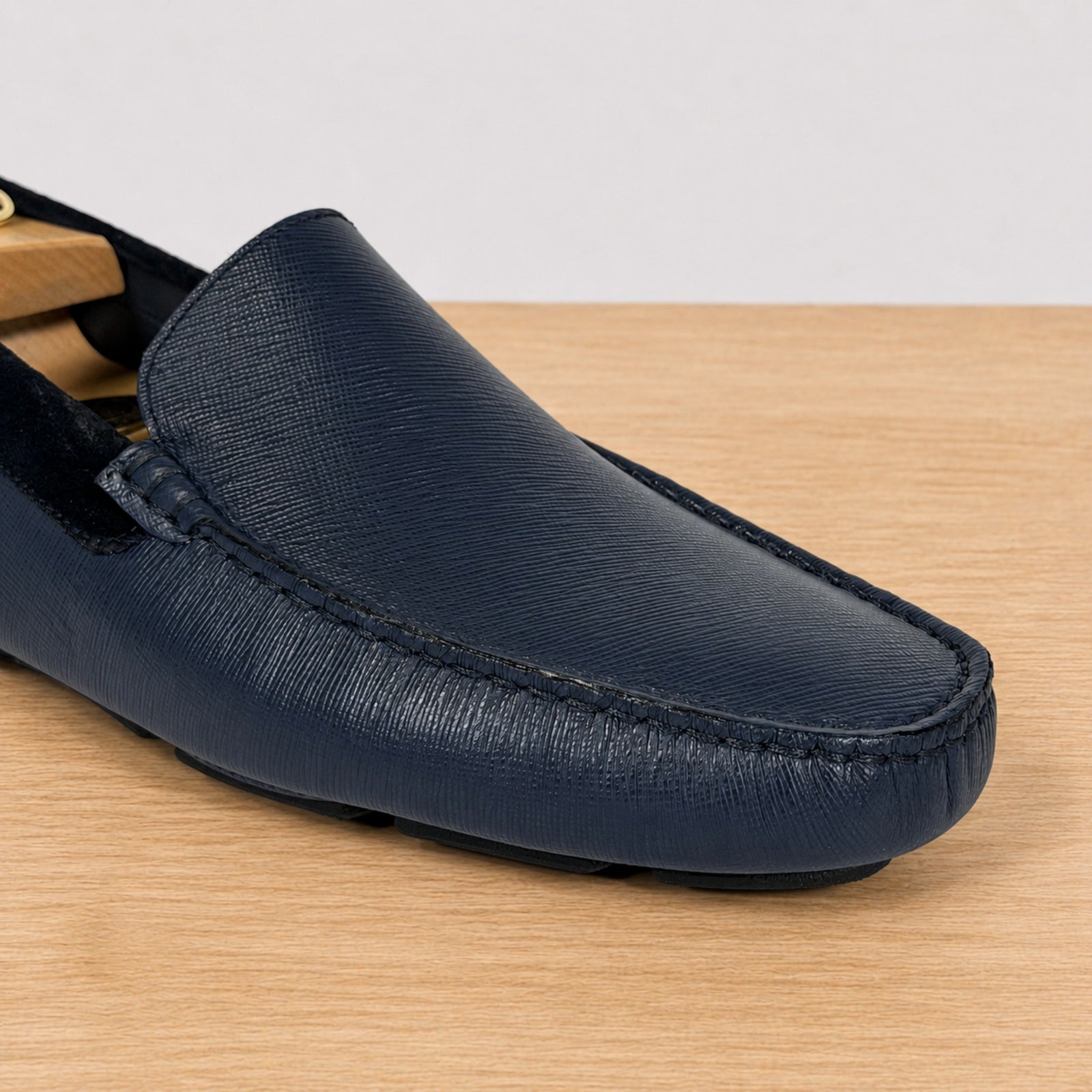 A close-up of a blue leather loafer with a textured grain finish, resting on a light wood surface against a neutral light gray background.