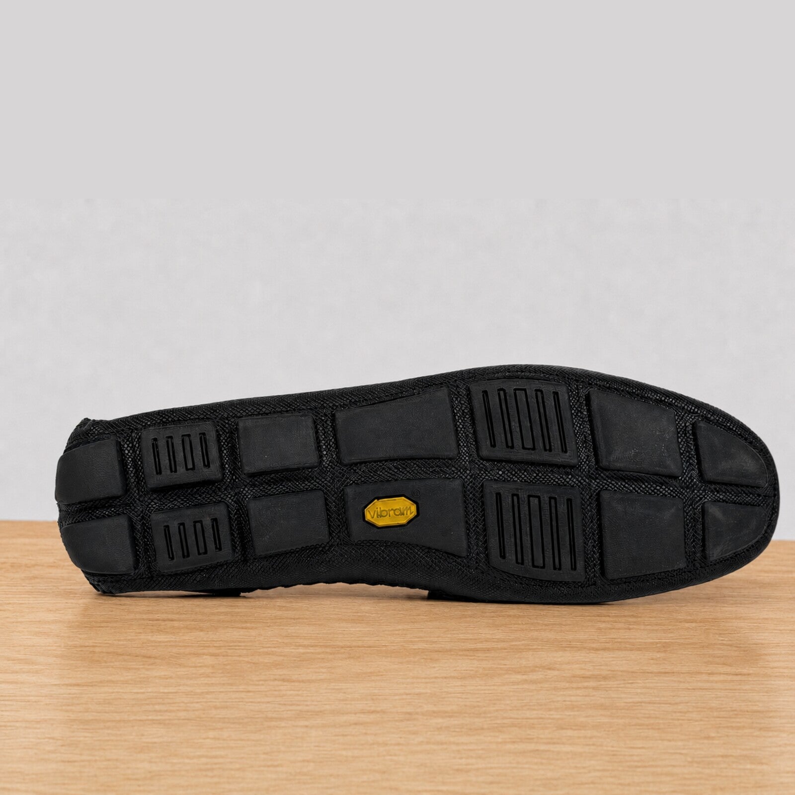 The black rubber outsole of a shoe, showing a grid tread pattern and a small yellow Vibram logo, resting on a light wood surface against a neutral gray background.