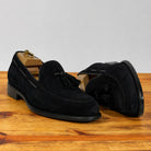 A pair of black suede tassel loafer shown in profile on a wooden surface against a light gray background.