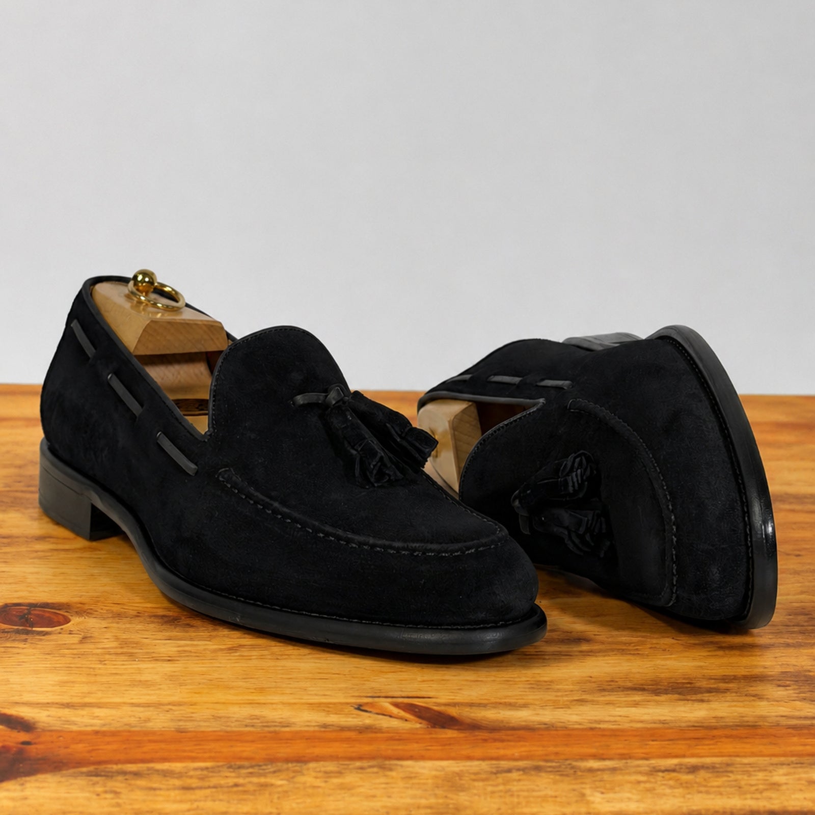 A pair of black suede tassel loafer shown in profile on a wooden surface against a light gray background.