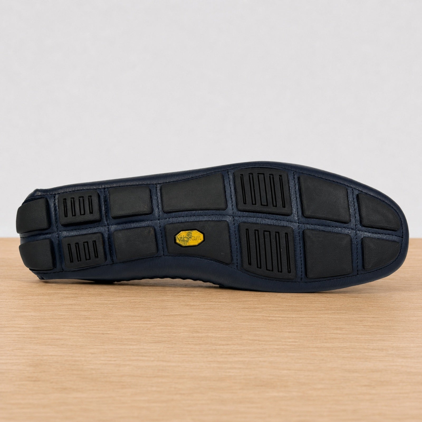 The outsole of a blue shoe resting on a light wood surface against a white background. The sole features a black tread pattern with a small yellow Vibram logo in the center.