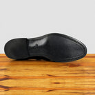 The outsole of a black leather dress shoe, showing the toe cap, stitching, and heel, resting on a wooden surface against a light gray background.