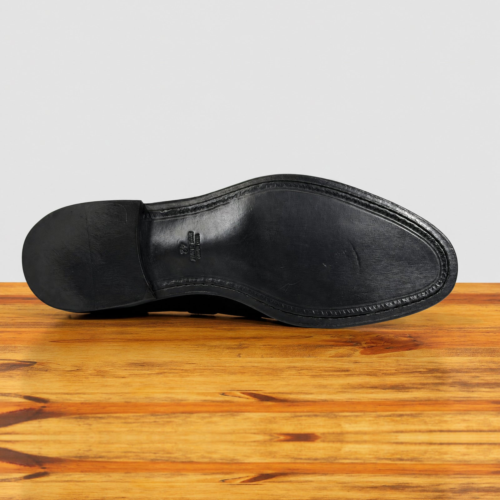 The outsole of a black leather dress shoe, showing the toe cap, stitching, and heel, resting on a wooden surface against a light gray background.