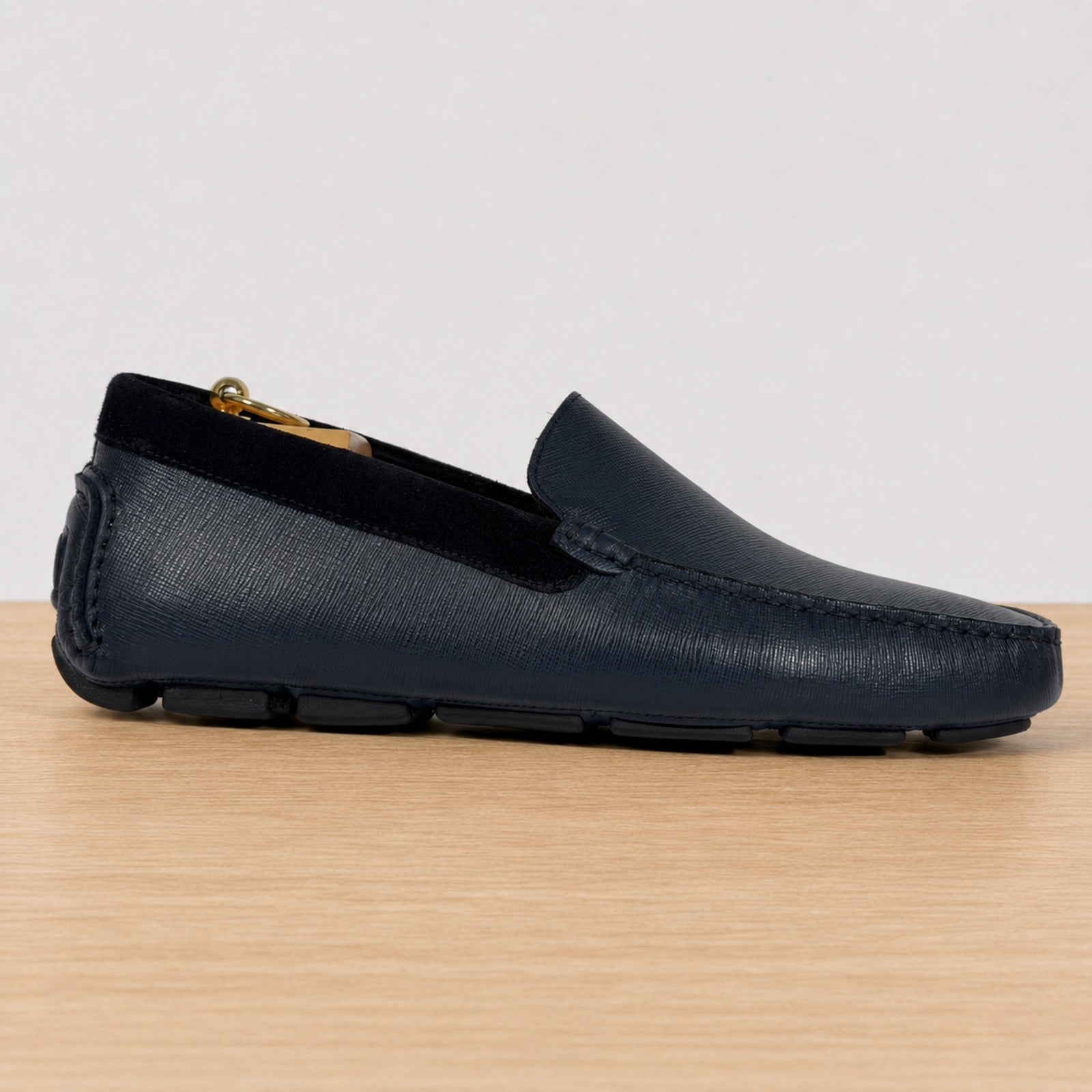A single  blue leather loafer with a textured grain finish, black velvet trim, shown in profile on a light wood surface against a neutral light gray background.
