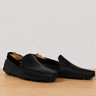 A pair of black leather driving loafers with a textured grain upper, displayed on a light wood surface against a neutral gray background.