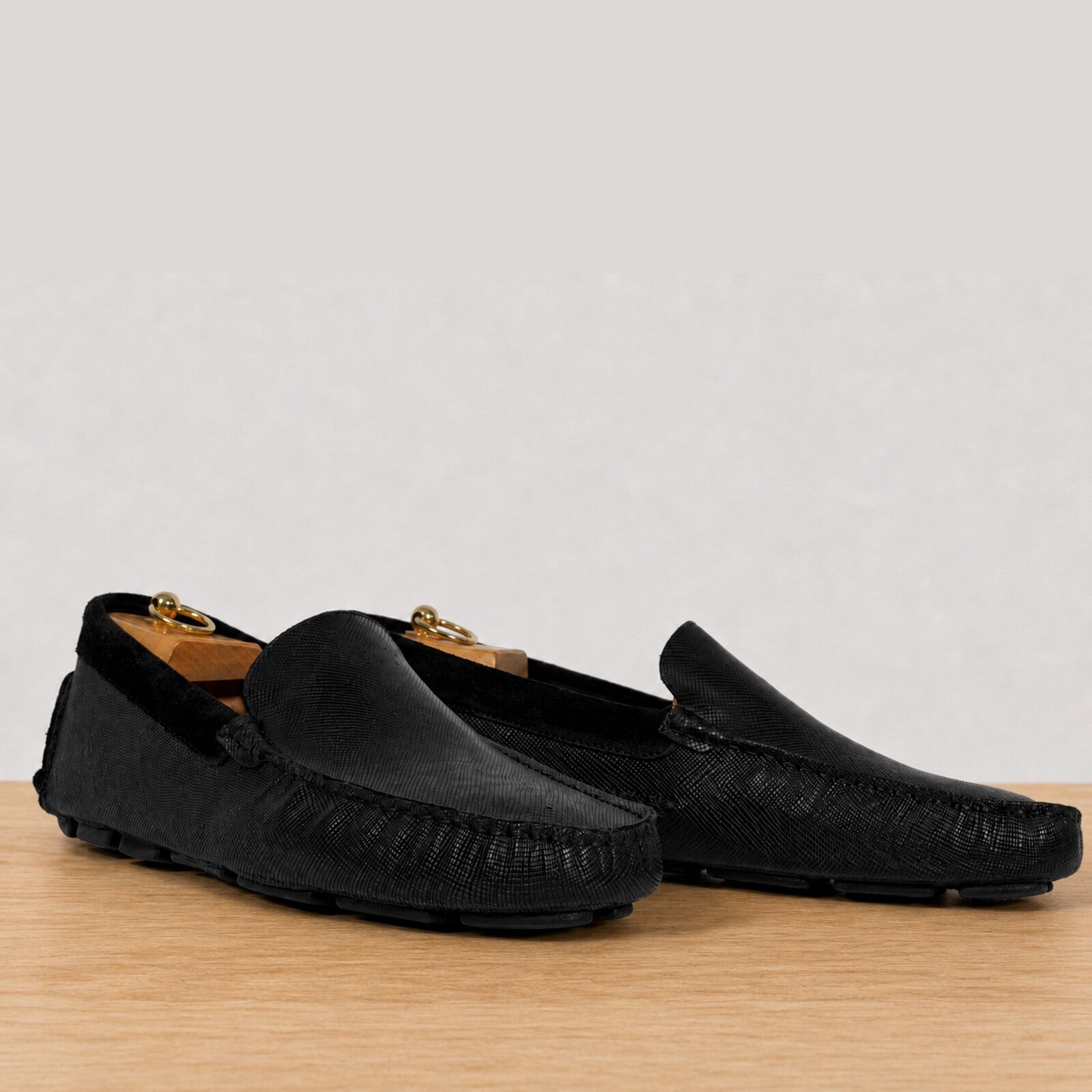 A pair of black leather driving loafers with a textured grain upper, displayed on a light wood surface against a neutral gray background.