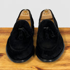 A pair of black suede tassel loafer shown in profile on a wooden surface against a light gray background.