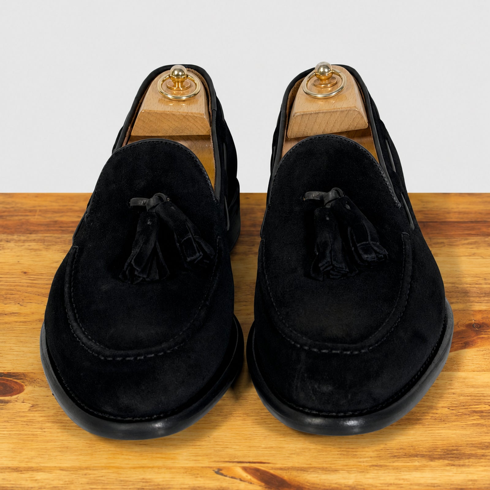 A pair of black suede tassel loafer shown in profile on a wooden surface against a light gray background.