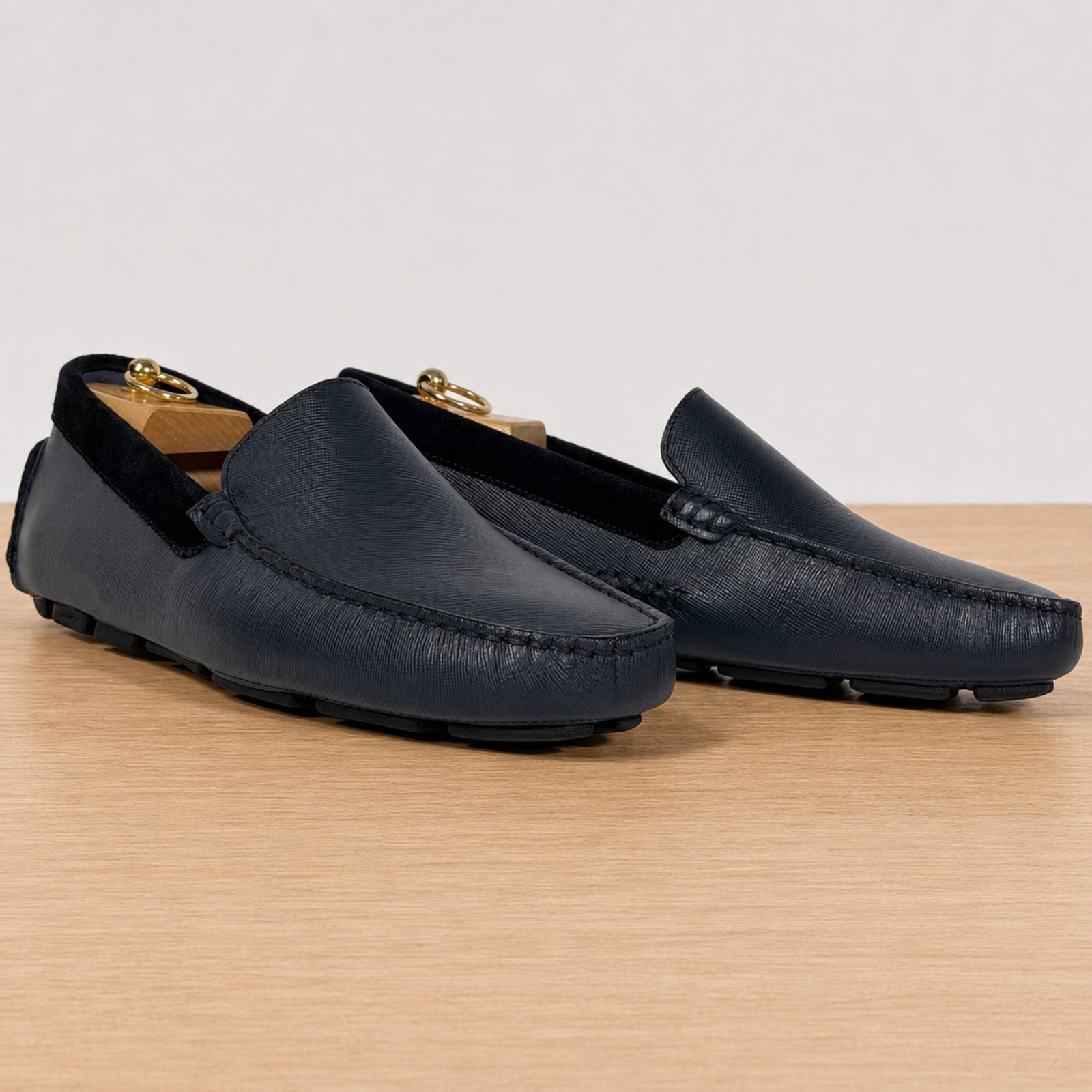 A pair of  blue leather driving loafers with black velvet trim, displayed on a light wood surface against a neutral light gray background.