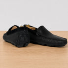 A pair of black leather moccasin-style driving loafers with textured detailing, displayed on a light wood surface against a neutral light gray background.