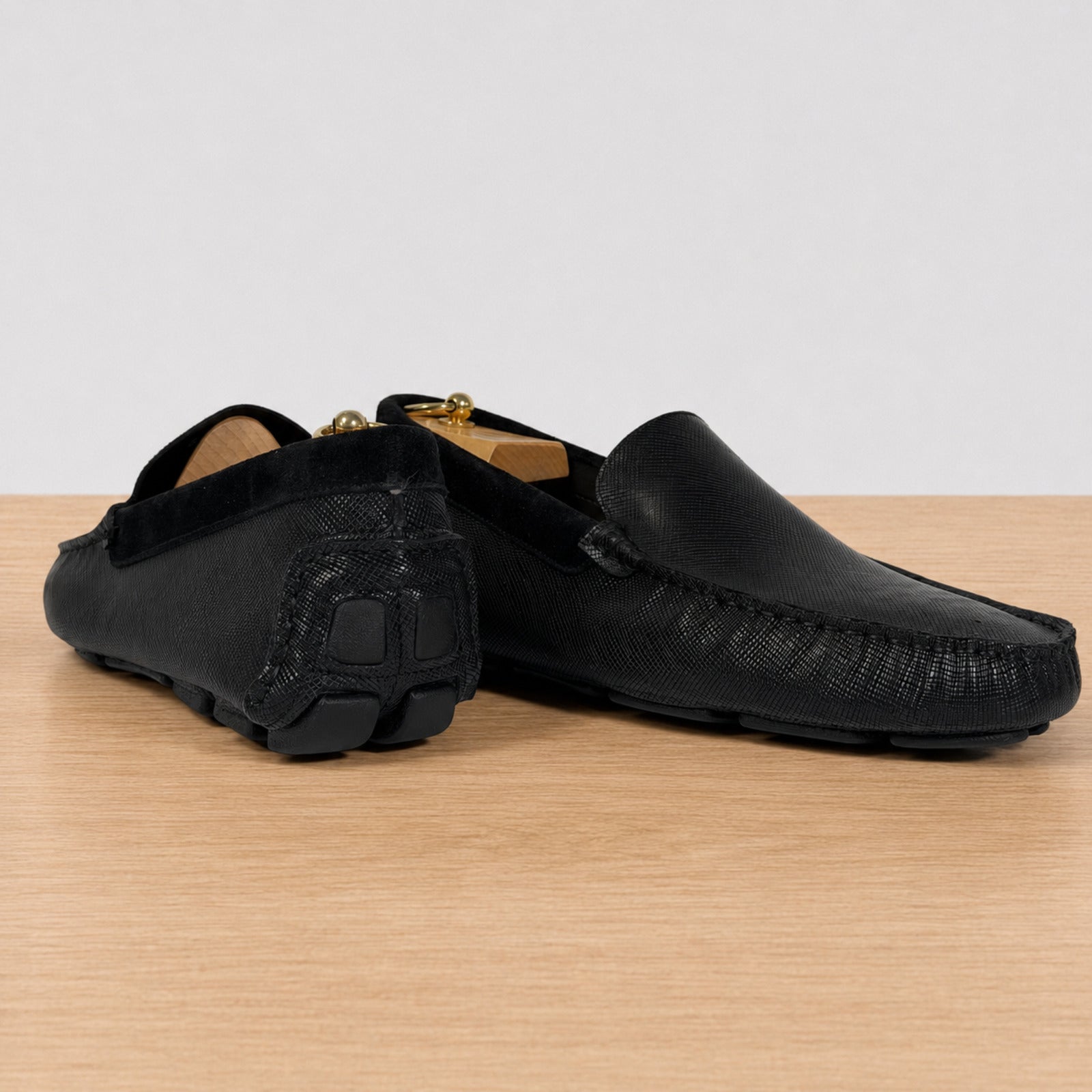 A pair of black leather moccasin-style driving loafers with textured detailing, displayed on a light wood surface against a neutral light gray background.