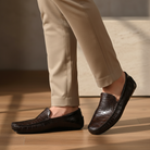 Brown leather loafers worn with beige pants on a wooden floor.