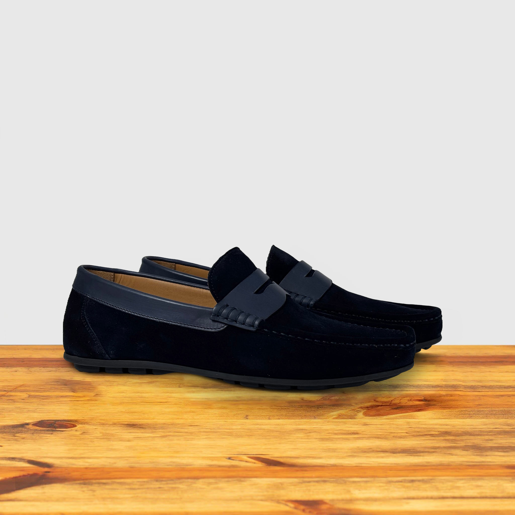 Pair of navy blue color suede penny loafer with a  strap, displayed on a wooden surface.