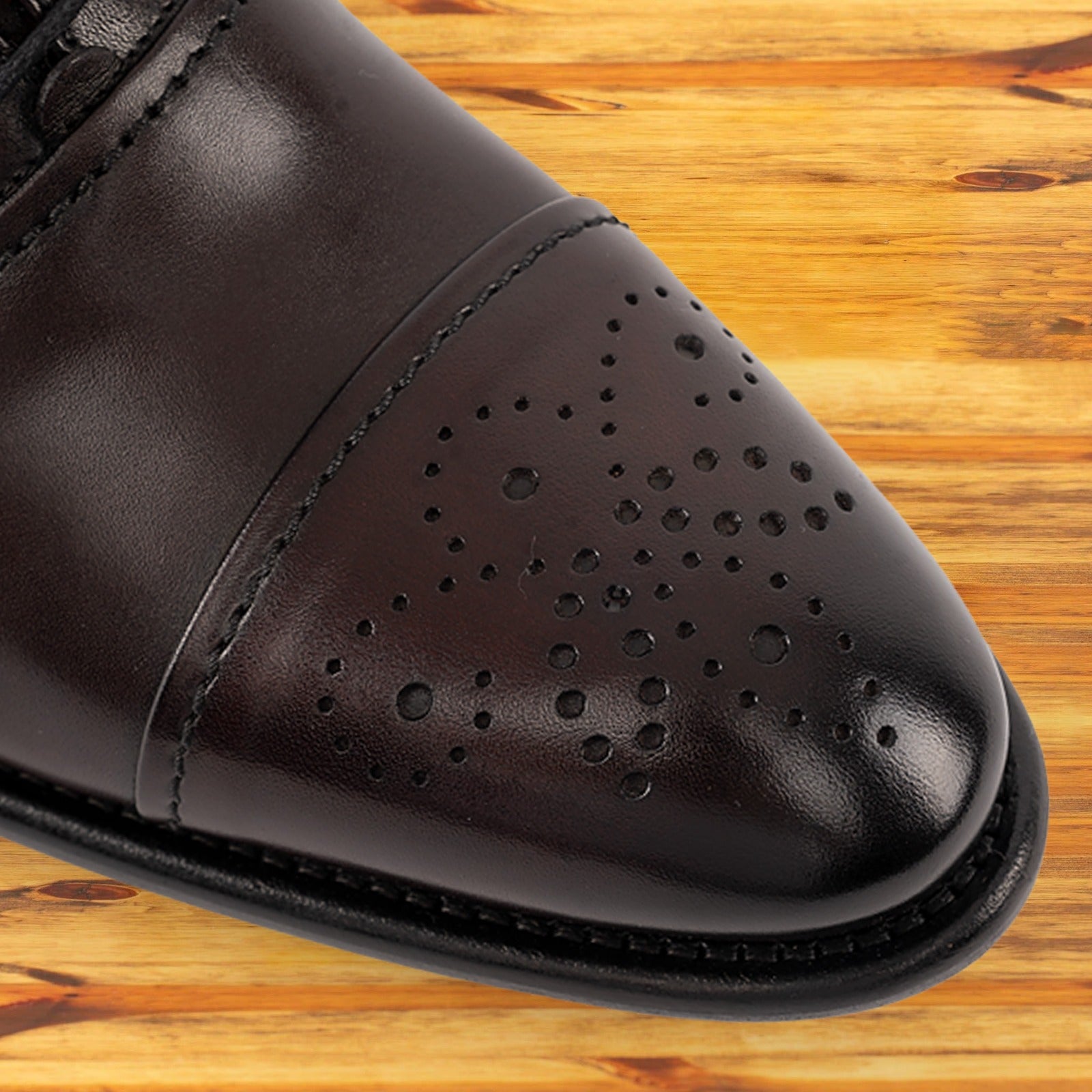 Close-up of a burgundy leather dress shoe toe featuring decorative perforations and stitching, resting on a wooden surface.