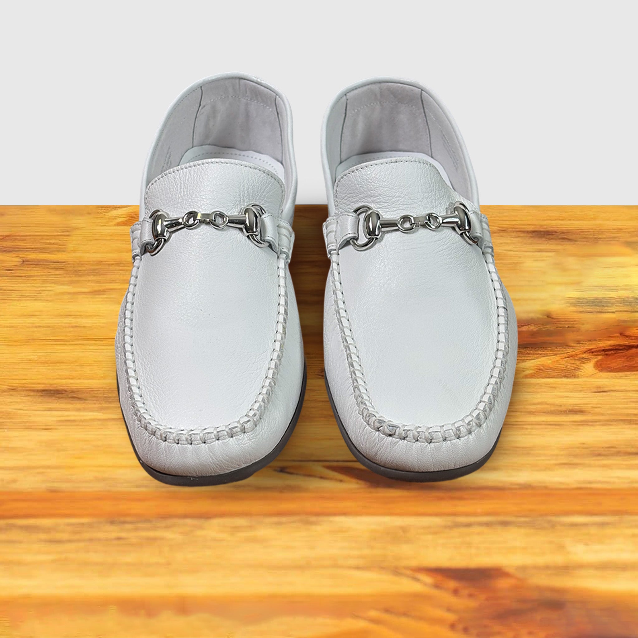 Pair of White loafer shoe with silver buckle on a wooden surface with a light gray background