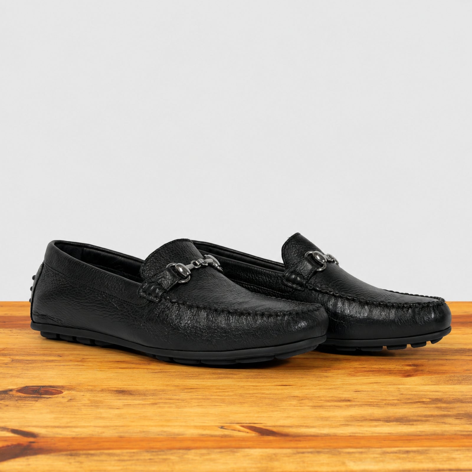 A pair of black leather driving loafers with a textured finish and a decorative metal buckle detail on the vamp, displayed on a wooden surface against a light gray background.