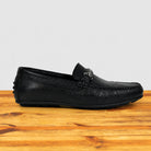 A single black pebbled leather loafer with a decorative silver-tone metal buckle detail on the vamp, shown in profile on a wooden surface against a light gray background.