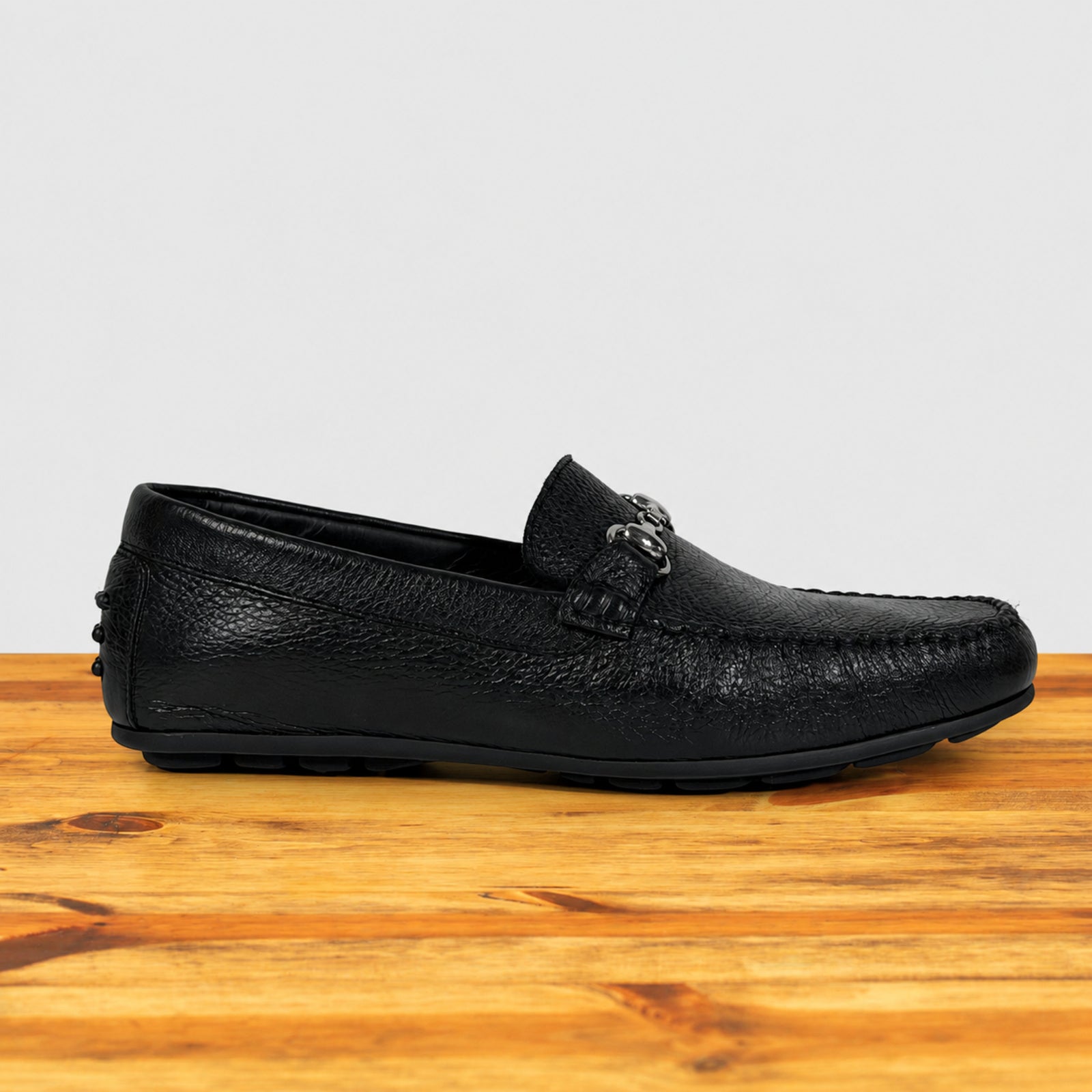 A single black pebbled leather loafer with a decorative silver-tone metal buckle detail on the vamp, shown in profile on a wooden surface against a light gray background.