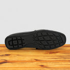 A black leather shoe shown from the bottom, displaying its textured treaded rubber sole, resting on a wooden surface against a light gray background.