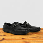 A pair of black leather loafers with a textured grain finish, displayed on a wooden surface against a light gray background.