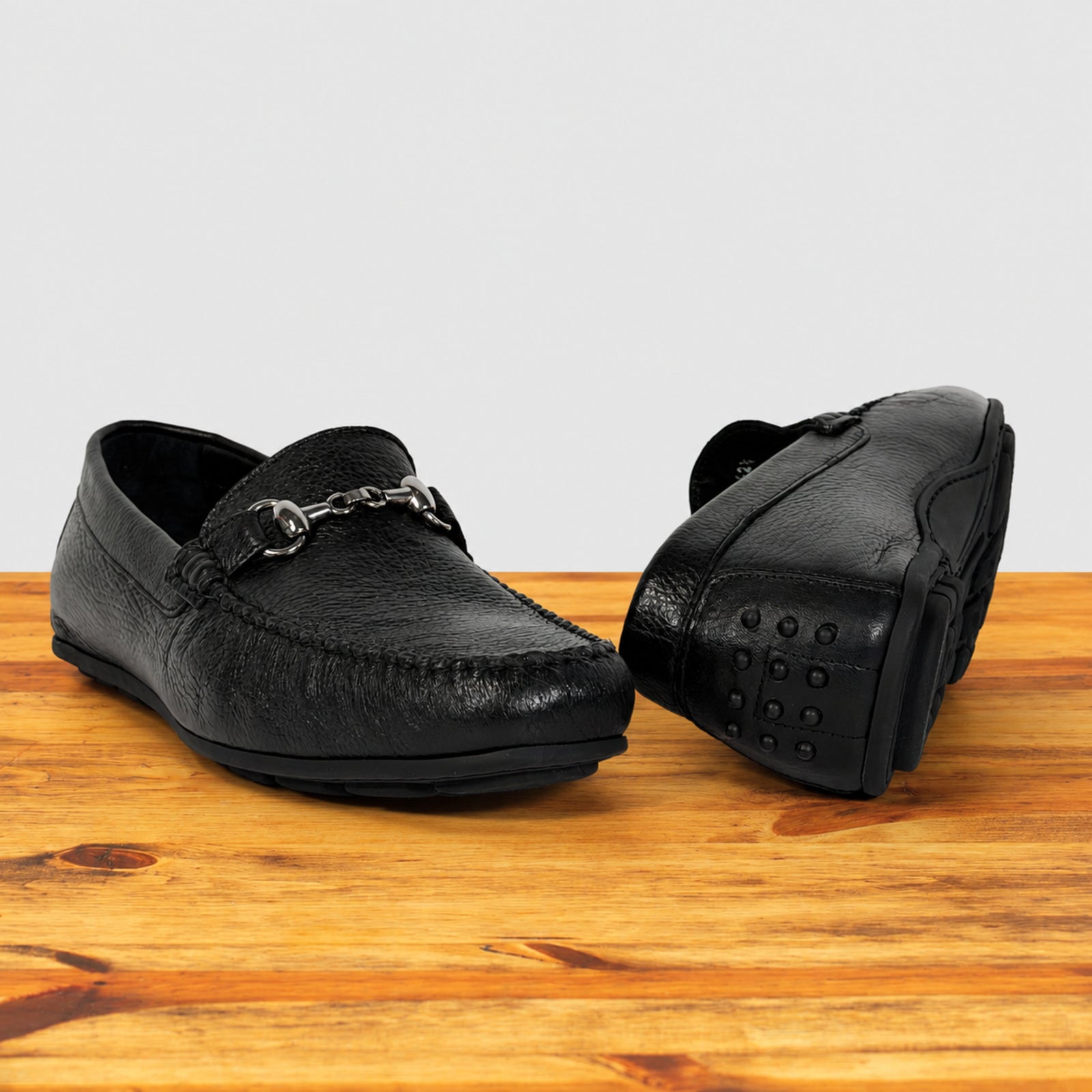 A pair of black leather driving loafers, displayed on a wooden surface against a light gray background.