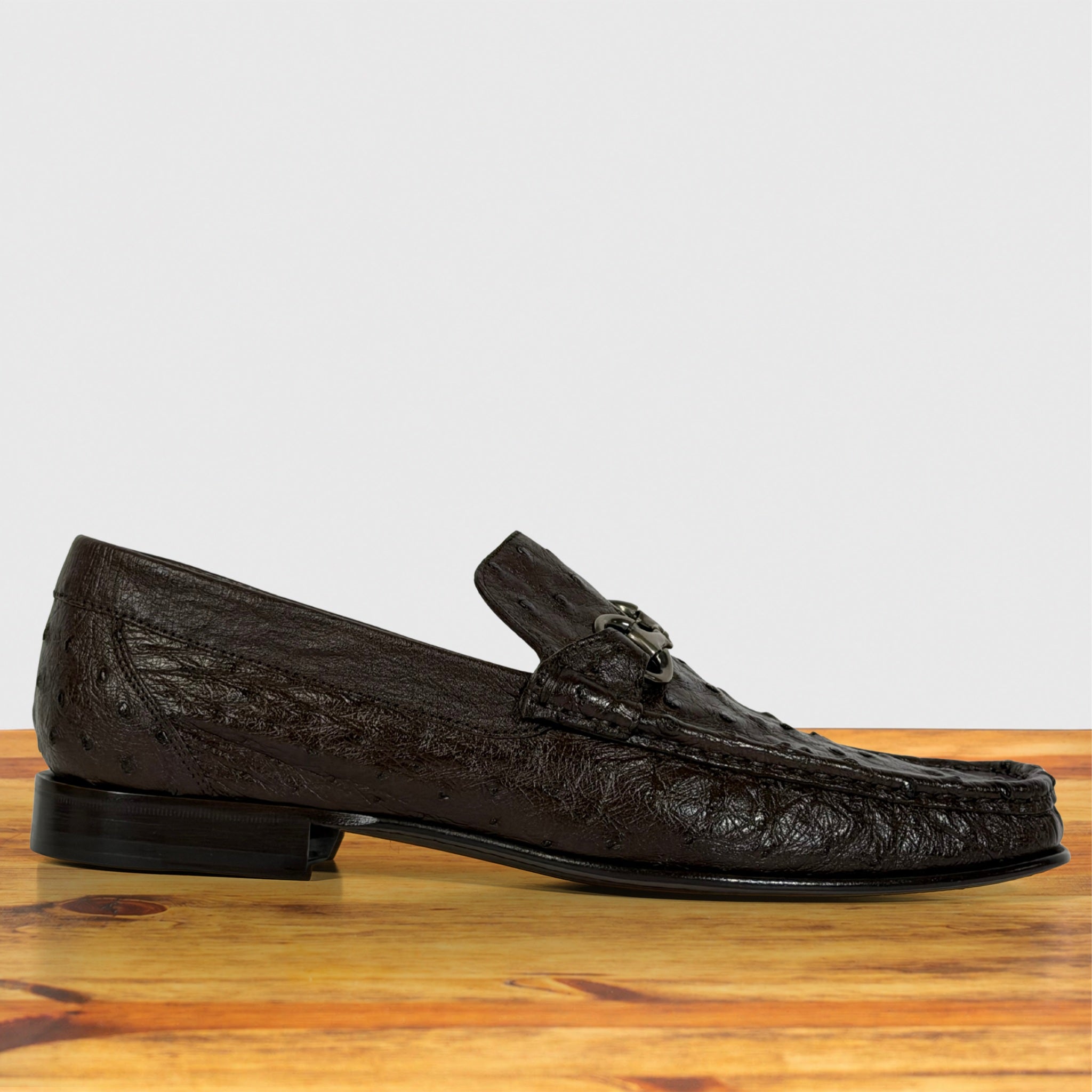A single dark brown textured leather loafer with a decorative strap and small metallic buckle, displayed on a wooden surface against a light gray background.