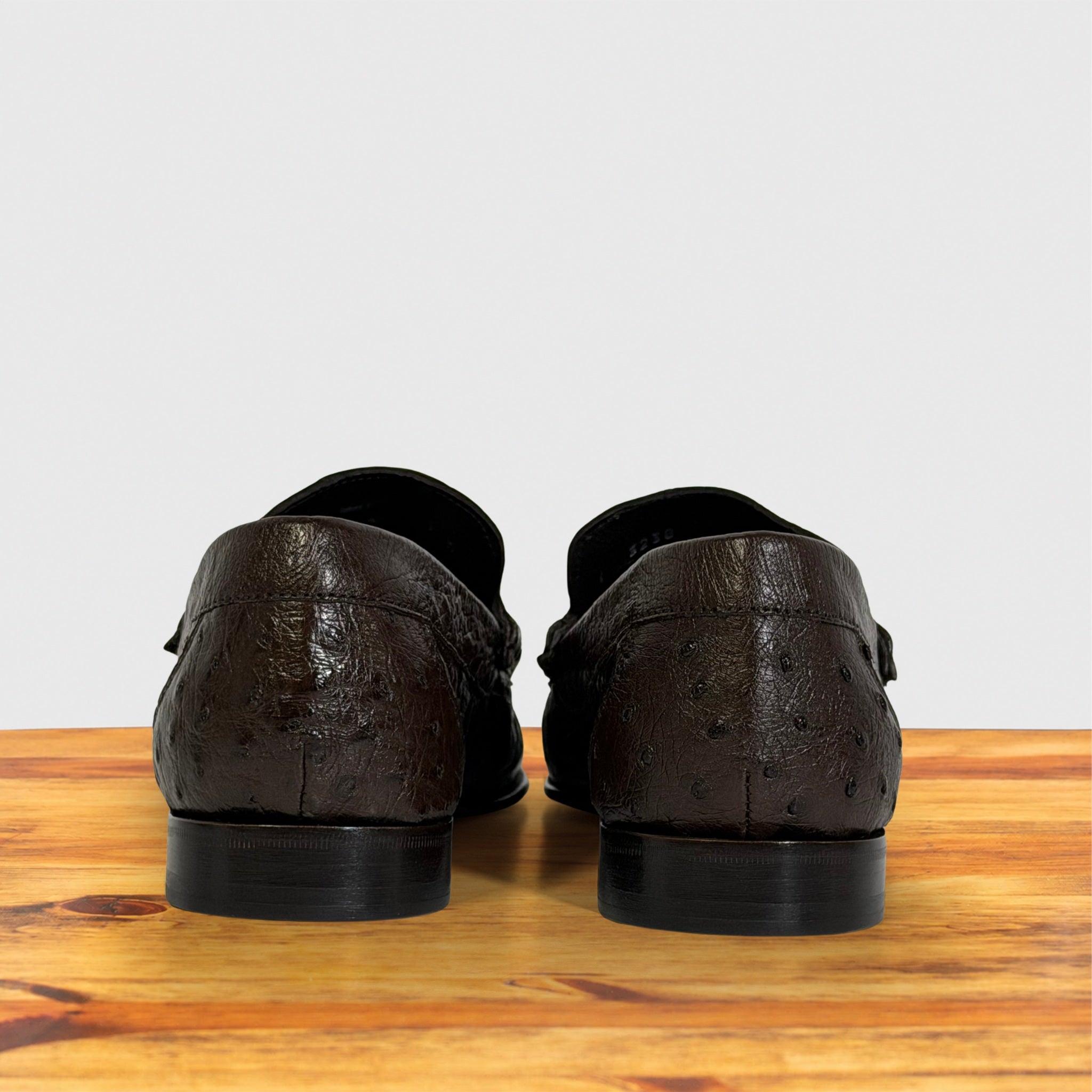 A pair of dark brown textured leather loafers shown from the back, resting on a wooden surface against a light gray background.