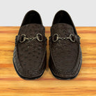 A pair of dark brown ostrich-textured leather loafers , displayed on a wooden surface against a light gray background.